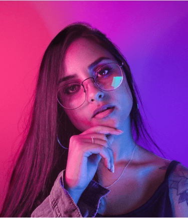 A young woman with long hair and glasses posing under vibrant pink and blue neon studio lighting.