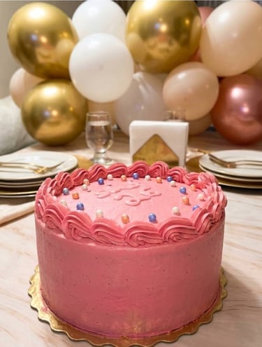 pastel de cumpleaños color rosa y de fondo una decoración de globos en dorado.