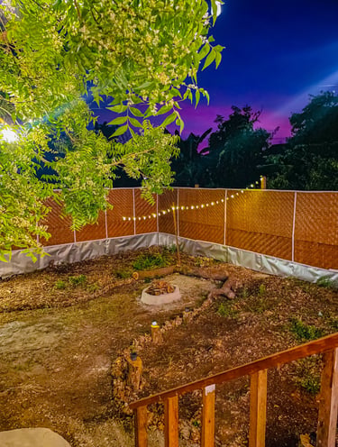 Backyard garden with a stone fire pit, bamboo fencing, and string lights under a purple twilight sky.