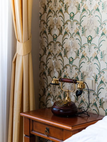 Vintage rotary phone on a wooden nightstand next to floral wallpaper and yellow curtains.