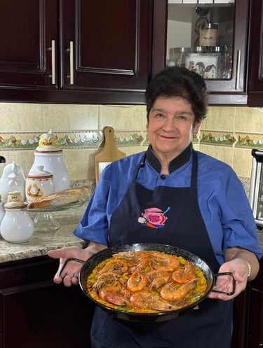 A smiling chef in a blue shirt holding a fresh seafood paella with large shrimp in a traditional pan.
