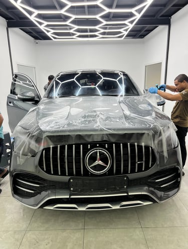 A technician applies clear paint protection film to a grey Mercedes-Benz SUV hood in a modern auto detailing shop.