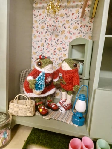 a couple of little knitted frogs wearing Christmas outfits in a doll house