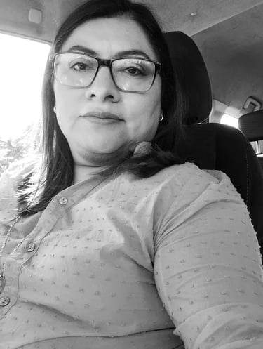 A black and white portrait of a woman wearing glasses and a polka dot shirt sitting inside a car.