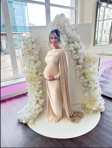 A pregnant woman in a beige maternity gown poses by a white rose flower arch at a baby shower.