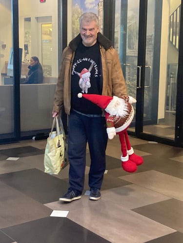 Willem in kersttrui met een kerstman en boodschappentas, lachend in de gang van een gebouw