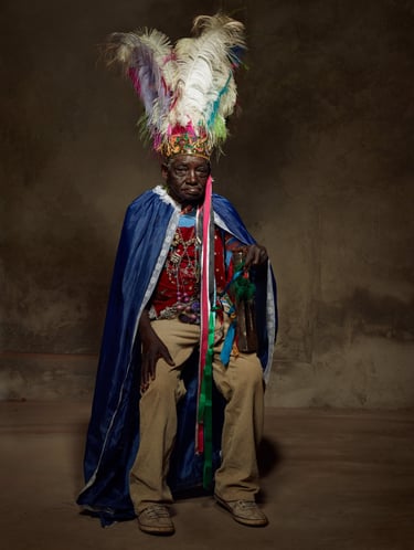 An old black man in a ritualistic gound with a feathered headdress, cape in a rugged cement backgrou