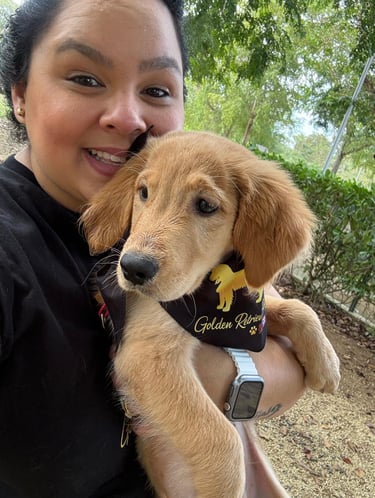 A smiling woman holding a fluffy Golden Retriever puppy wearing a black bandana outdoors.