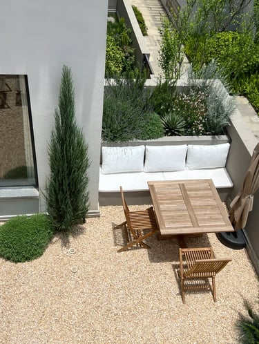 Simple low-maintenance garden corner with gravel ground, wooden table, outdoor chairs, white walls a