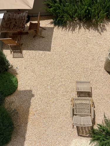 Minimal outdoor seating area with gravel ground, wooden furniture, umbrella shade and green shrubs f