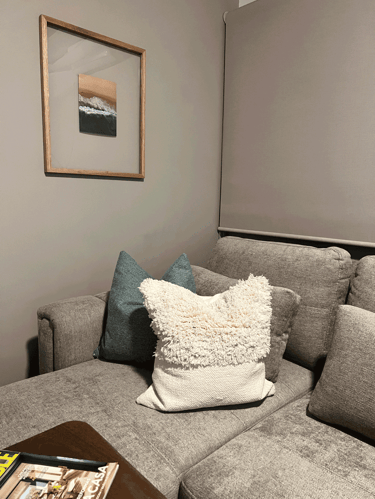 Rincón de sala con cojines de textura, cortinas enrollables y cuadro de paisaje costero | KARÄNA