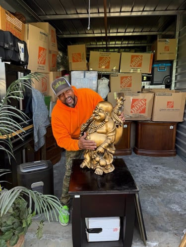 A man in an orange hoodie holds a large golden Buddha statue in a storage unit filled with moving boxes.