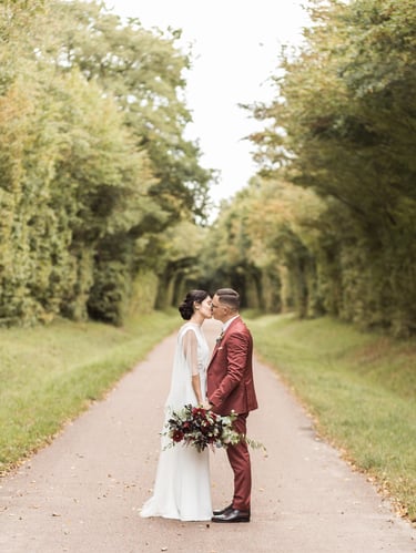 A bride in a white lace dress and a groom in a red suit kissing on a tree-lined road.