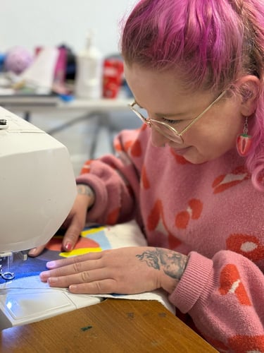 Harriet Mee, a woman with pink hair and glasses is sewing a picture