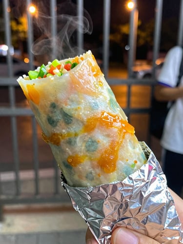 Vibrant food photography, a steaming roll wrapped in foil held by a student near a hostel gate at night under warm streetlights, close-up macro, shallow depth of field.