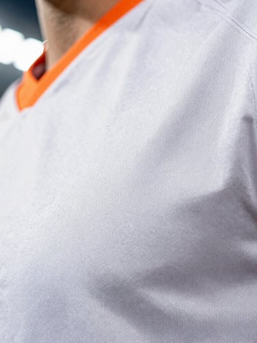 Crisp close-up of high-performance technical fabric jersey stitching with subtle orange accents, bright stadium lighting, sharp focus.