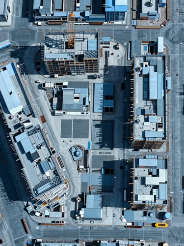 Top-down orthomosaic map of an active commercial construction site, sharp technical photography, bright midday sun, high-contrast blue and slate tones, CAD grid overlay style.