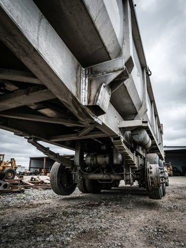 Low-angle dramatic shot of a heavy-duty aluminum dump trailer chassis under overcast industrial skies, highlighting metallic weld textures, 35mm.