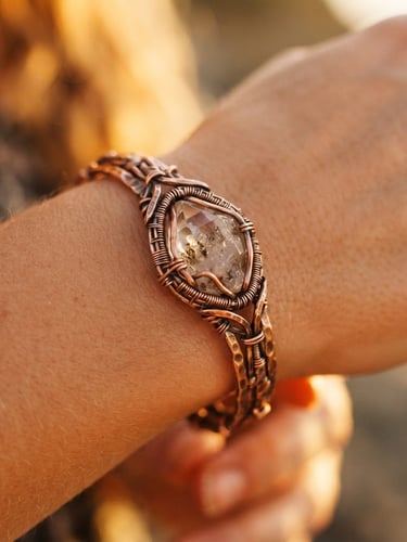 Close-up of a handcrafted copper bracelet with a raw crystal, resting on warm, bare skin, illuminated by golden-hour sunlight. Shallow depth of field, highlighting the hammered texture of the metal.