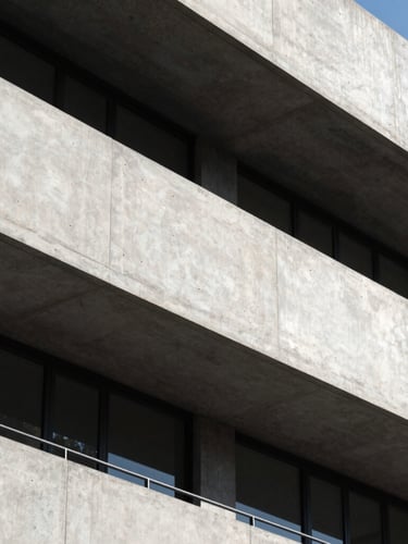 High-contrast architectural crop of a modern concrete and steel facade, sharp geometric shadows under bright daylight, clean lines, minimalist composition.
