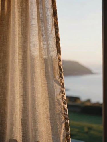 A close-up portrait of textured linen curtains blowing in a soft breeze through an open window overlooking a misty Welsh coastline at dawn, warm morning light, shallow depth of field.