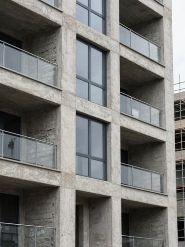 Close standoff shot of a concrete and glass residential facade, framing reveals exposed structural columns and rough masonry infill, natural overcast daylight, scaffolding partially visible on the right edge