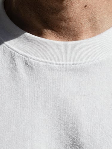 Extreme close-up of a thick ribbed collar and dense cotton weave of a white t-shirt, high-contrast desaturated tone, sharp shadows, raw industrial texture.