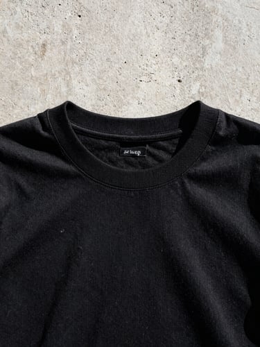 Low-angle close-up of a heavy-weight 250 GSM black cotton t-shirt showing the thick tight collar and dense fabric texture against a raw concrete Manila wall under harsh midday sunlight.