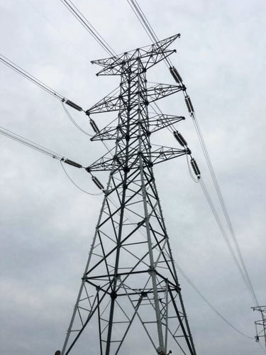 Wide environmental shot looking up along high-voltage transmission pylons receding into overcast sky, industrial steel lattice in sharp relief, photographed from below at architect's elevation angle, no people, developed-market landscape