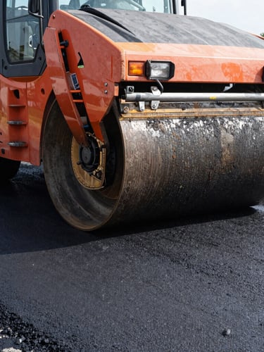 Low-angle heroic shot of a heavy asphalt roller on fresh textured black pavement, high-contrast morning light, safety orange accents on machinery, 35mm.