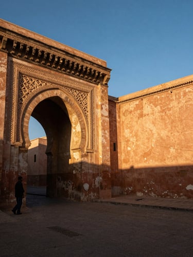 Wide panoramic low-angle view of a weathered stone archway in an ancient Moroccan medina, a lone male traveller silhouetted in the arch's shadow at far left, late afternoon golden light raking across carved plasterwork and terracotta walls, deep atmospheric shadows in the foreground, generous sky and warm stone fill the right two-thirds