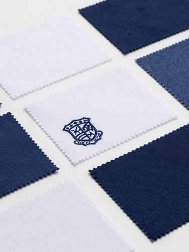 Close-up of various fabric swatches in different shades of blue and white, with one swatch featuring a small, finely embroidered school crest. Bright, clean studio lighting, sharp focus on textures.