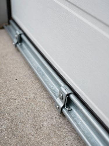 Extreme close-up of a freshly-installed garage door track and roller bracket, shot from low angle looking up the driveway, natural overcast daylight raking across the galvanized steel hardware, concrete driveway edge visible at bottom left, no people
