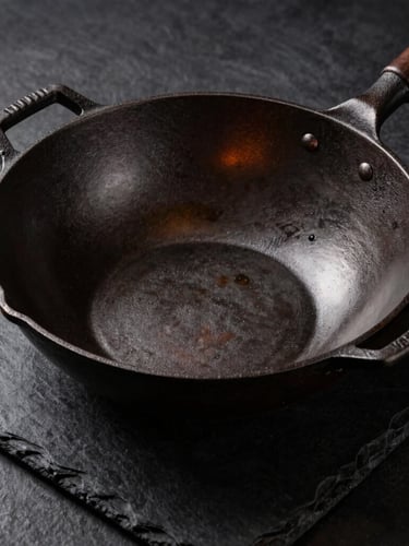 High-contrast editorial shot of an uncoated cast iron wok on a dark slate surface, showing raw iron texture, dramatic side lighting