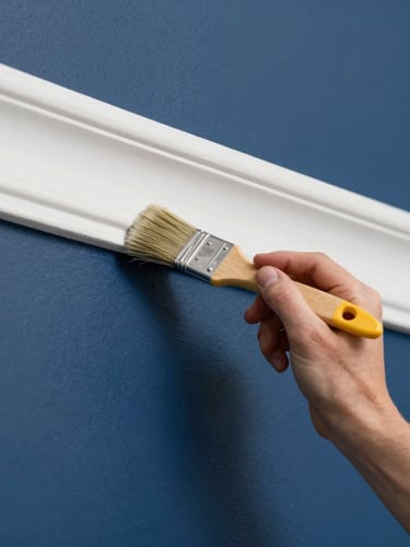 Close-up action shot of a professional painter's hand using a precision sash brush to cut a clean white trim line against a deep navy blue wall, bright daylight.
