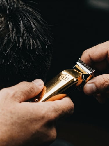 Close-up of a professional barber's hands using gold-accented clippers to sculpt a sharp fade, moody low-key lighting, deep black background, warm gold reflections