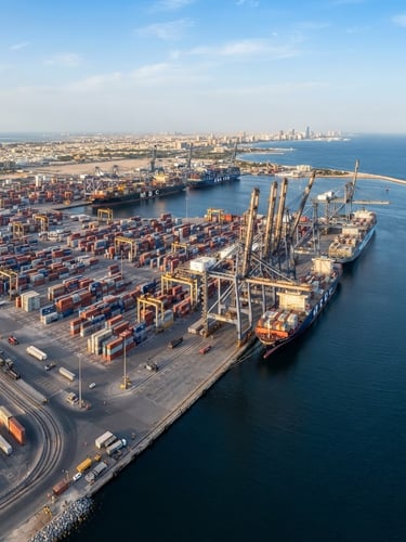 High-angle panoramic view of a deep-water shipping port in Sharjah, massive cargo containers stacked neatly under bright daylight, cool navy water, steel cranes reflecting gold highlights.