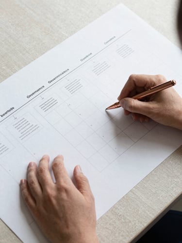 Overhead flat-lay of a large printed strategy roadmap canvas on a pale linen surface, hands annotating a governance matrix with a copper-tipped pen, soft diffused daylight from above, sparse and precise composition with visible grid lines and framework stages
