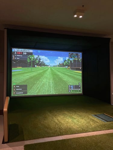 Wide environmental shot of a premium indoor golf simulator bay, glowing screen showing a virtual course fairway, moody low-key ambient lighting, deep charcoal tones, 35mm