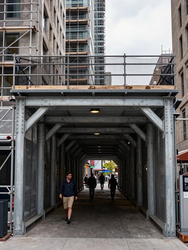 Panoramic eye-level view of a reinforced pedestrian tunnel installed beneath a high-rise facade under active renovation in downtown Montreal, overcast daylight, scaffolding and safety netting above, pedestrians walking through the corridor, load-bearing connection points visible on the steel uprights