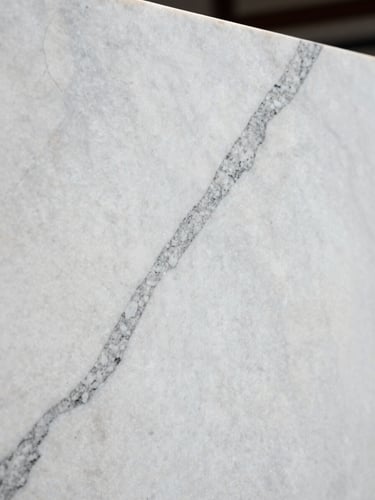 Side-angle close-up of a polished white Statuario marble slab under cool north-facing daylight, bold dark grey veining running diagonally across the frame, sharp grain texture visible at full resolution, no staging