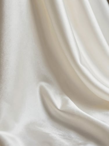 Macro studio photograph of heavy ivory bridal satin draped in elegant geometric folds, soft side-lighting highlighting the subtle sheen and luxurious weight of the weave.