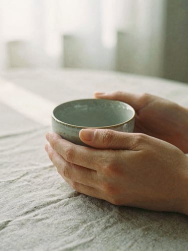Close-up of hands holding a warm ceramic cup on a textured linen tablecloth, soft morning sunlight filtering through a window, muted earth tones, 35mm film style