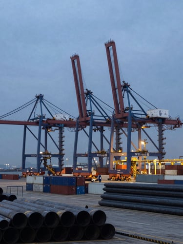 Wide environmental shot of a deep-water industrial port terminal at twilight, massive container cranes silhouetted against a cold overcast sky, active laydown yards with stacked steel pipes in the foreground.