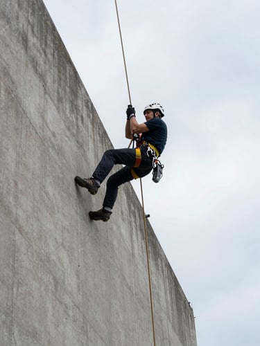 Wide shot from below looking up at a rope access technician in full harness and helmet descending a concrete industrial façade, safety rigging visible against overcast daylight sky, structural surface texture sharp in foreground