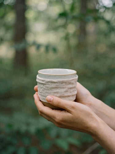 Soft diffused natural daylight filtering through forest leaves, a woman's hands holding a warm ceramic mug wrapped in soft linen, 35mm film style.