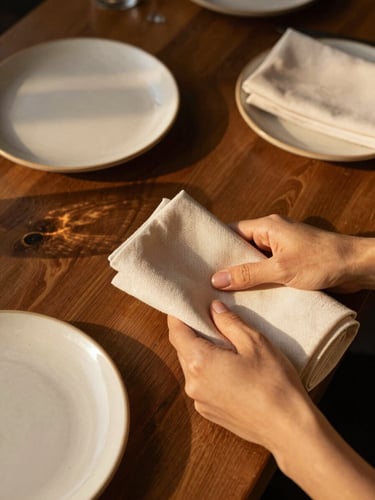 Close overhead view of a restaurant prep table, hands folding linen napkins beside ceramic plates, warm golden-hour light raking across the surface, dark wood grain visible underneath