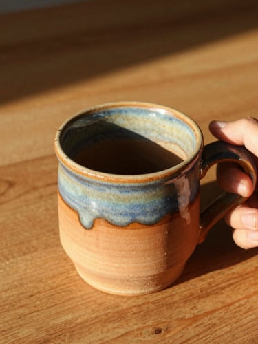 Overhead warm editorial shot of a hand holding a rustic terracotta-glazed mug, bright golden-hour sunlight casting soft shadows on a wooden table, 35mm