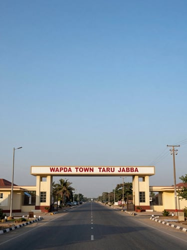 Wide panoramic view of WAPDA Town Taru Jabba main entrance gate and signage, straight paved road leading inward under clear blue sky, utility poles flanking both sides, late-morning daylight, framed to fill the right half of the composition with generous sky above