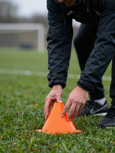 Tight action close-up of a football coach in a dark athletic jacket placing orange agility cones on a dew-covered grass pitch, overcast daylight, shallow depth of field, 35mm